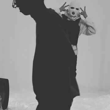 A grainy black-and-white frame of two figures, one masked, flashing a peace sign