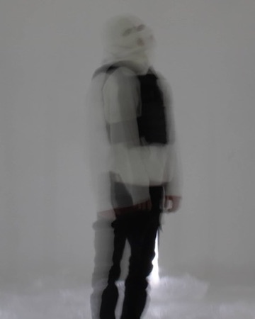 A long-exposure blur of a hooded figure against a white wall