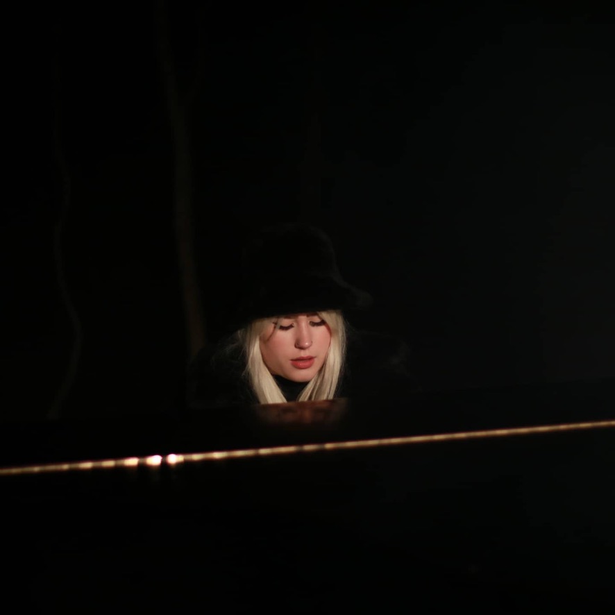 A figure in a black hat at a grand piano, lit by a single warm light